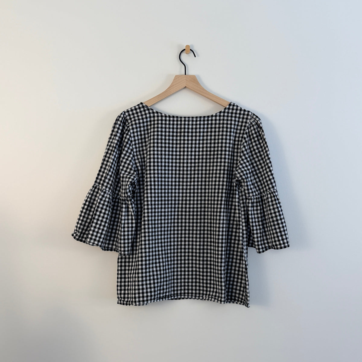 A black and white gingham patterned blouse hanging on a wooden hanger against a plain white wall.